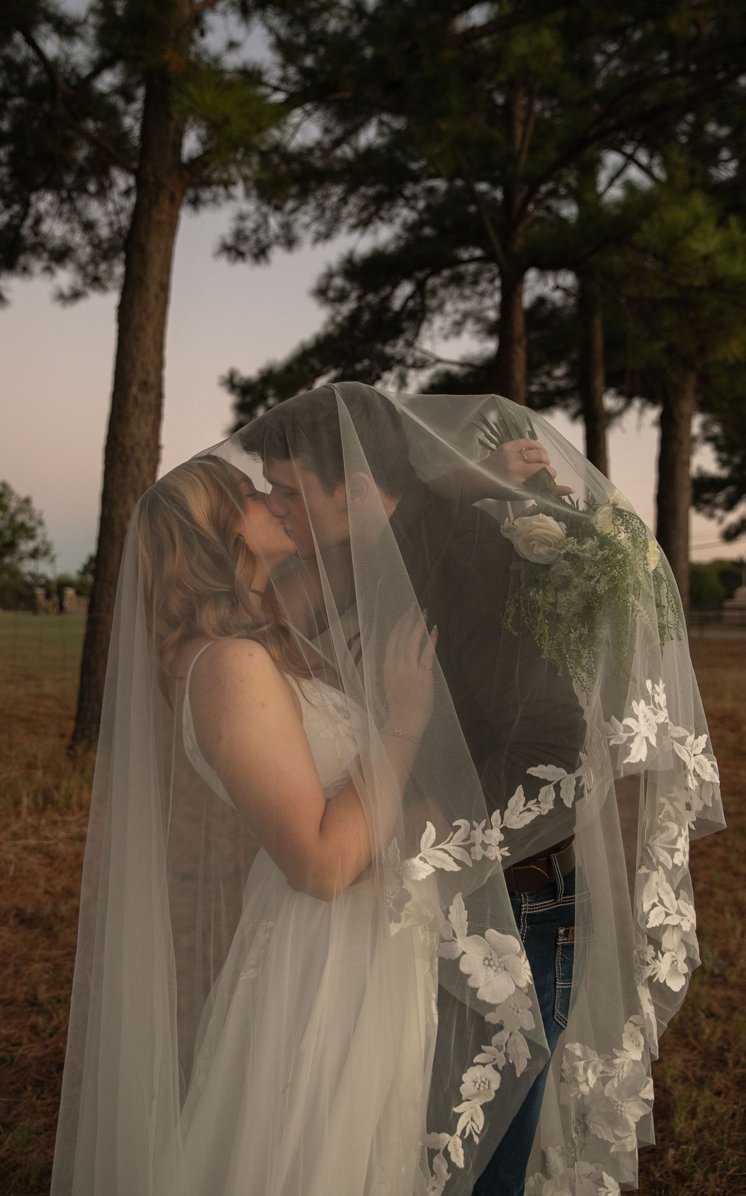 Bride and groom share a romantic kiss hidden beneath a flowing lace wedding veil outdoors among pine trees.