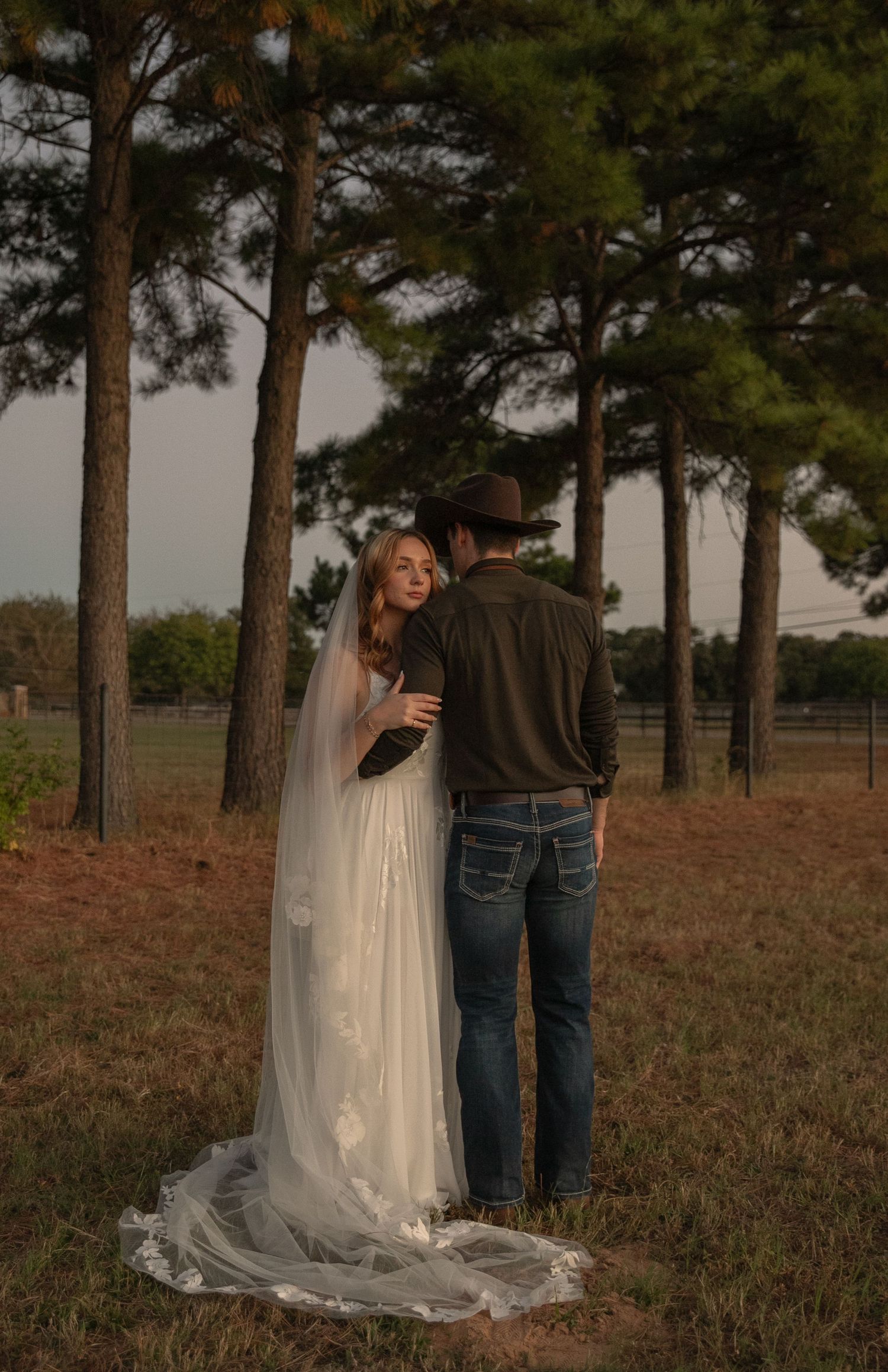 Bride in white gown and veil embracing groom in cowboy hat and jeans among tall pine trees at sunset.