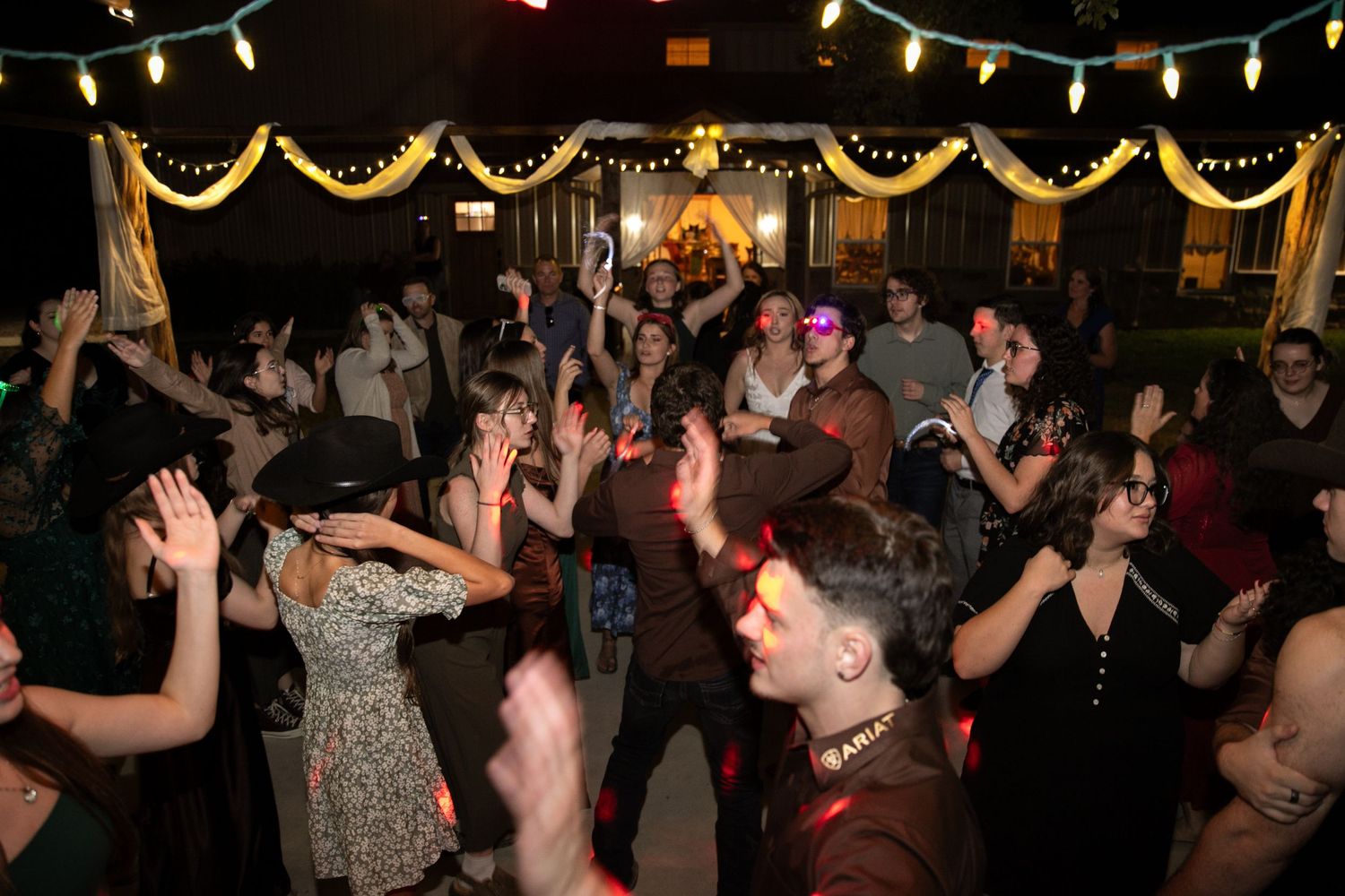 Energetic wedding reception dance floor filled with guests dancing under warm string lights at night.