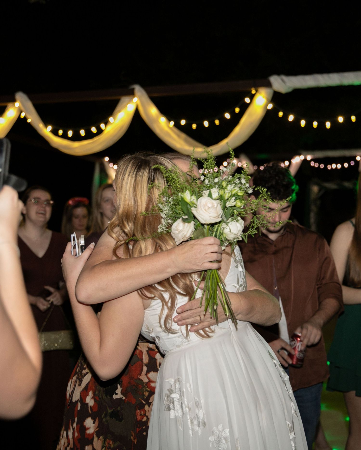 Bride holding white floral bouquet sharing a hug with a guest on the wedding reception dance floor.