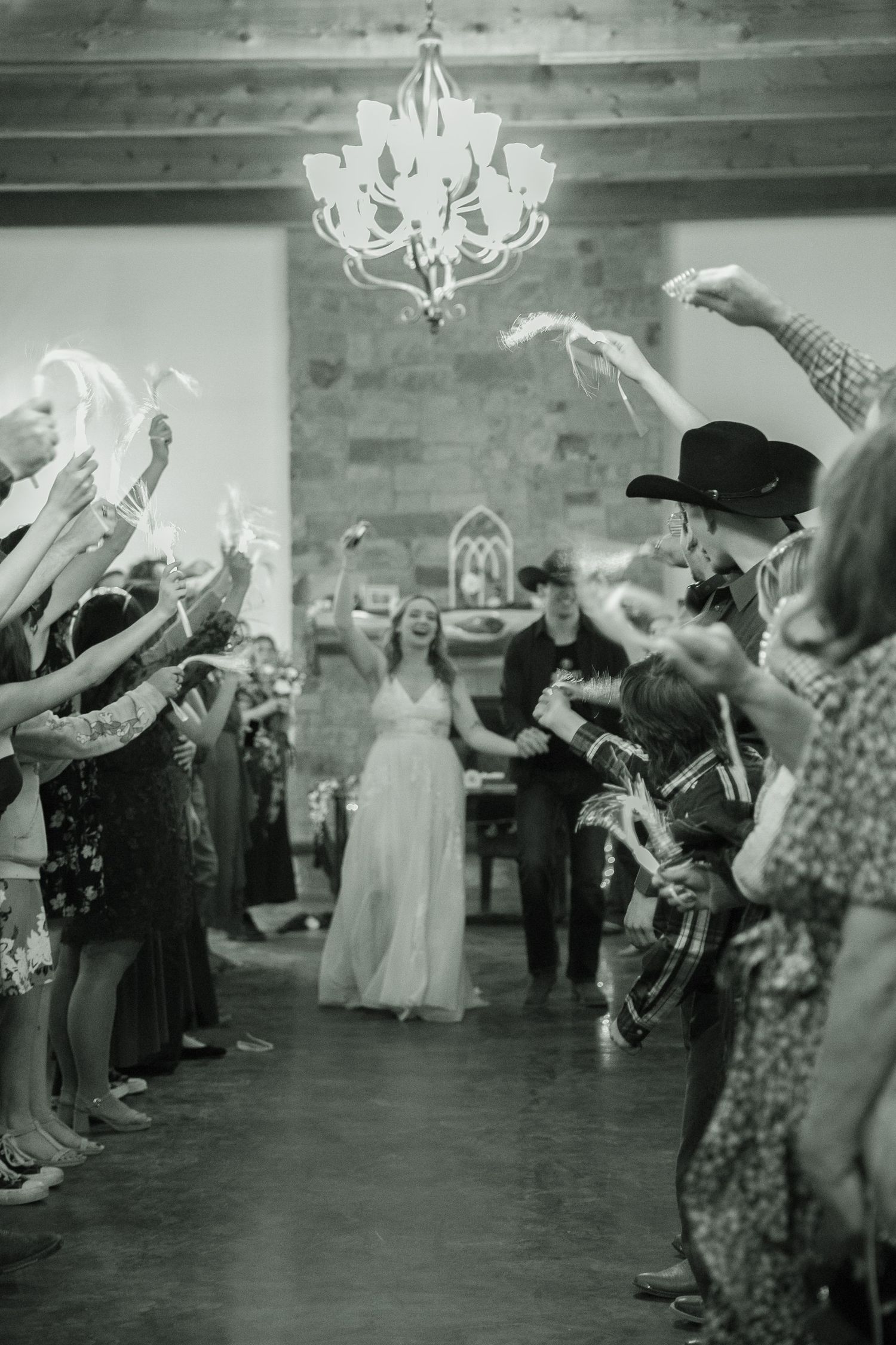 Black and white photo of bride and groom exiting through a celebratory sendoff line under a chandelier.