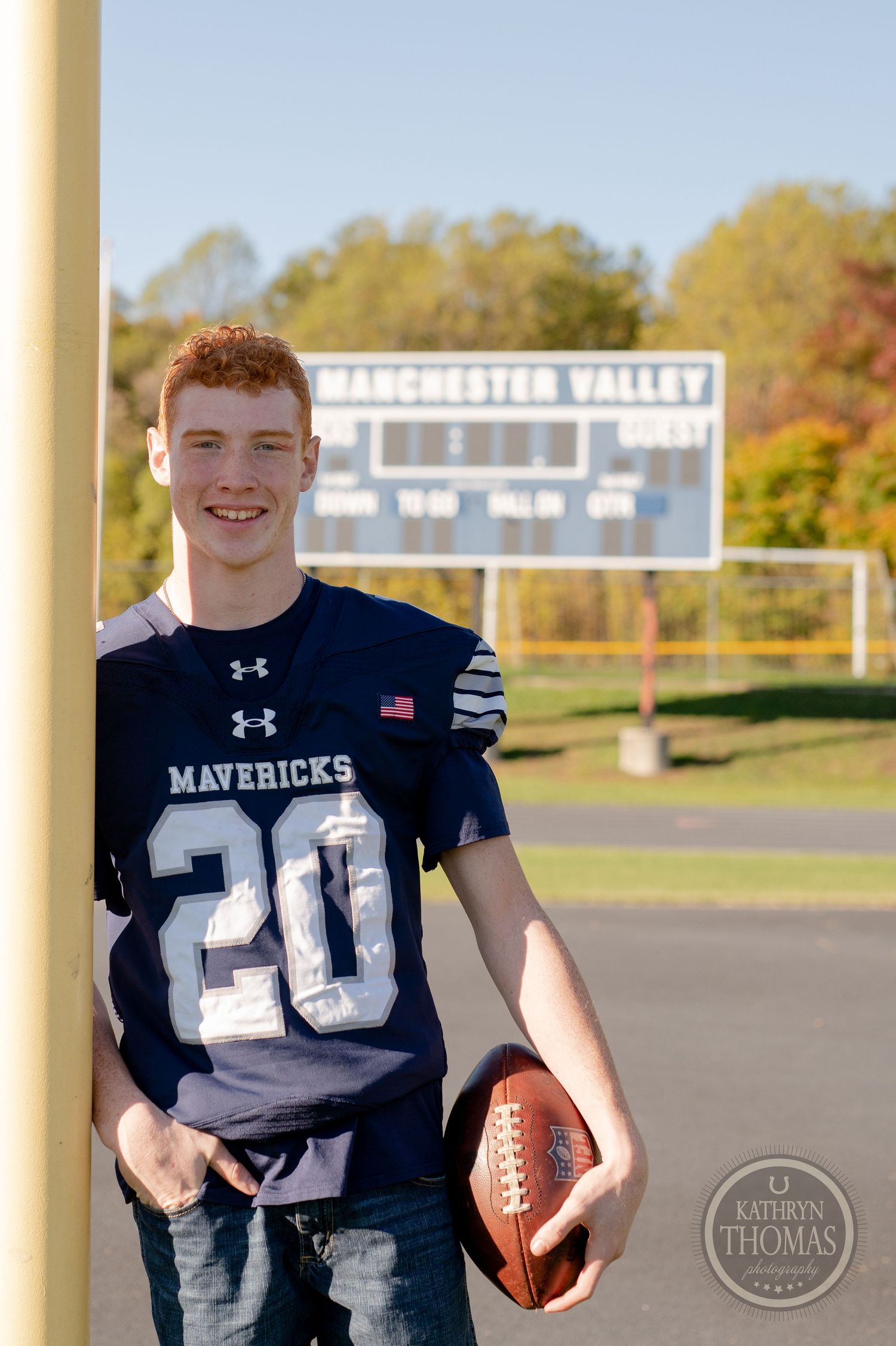 A high school football player in jersey number 20 poses on the field with the Manchester Valley scoreboard in the background.