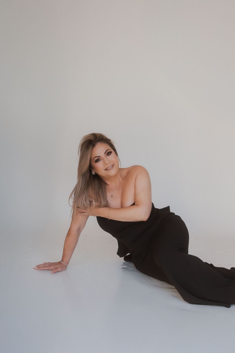Woman in strapless black gown poses artistically on white studio floor.