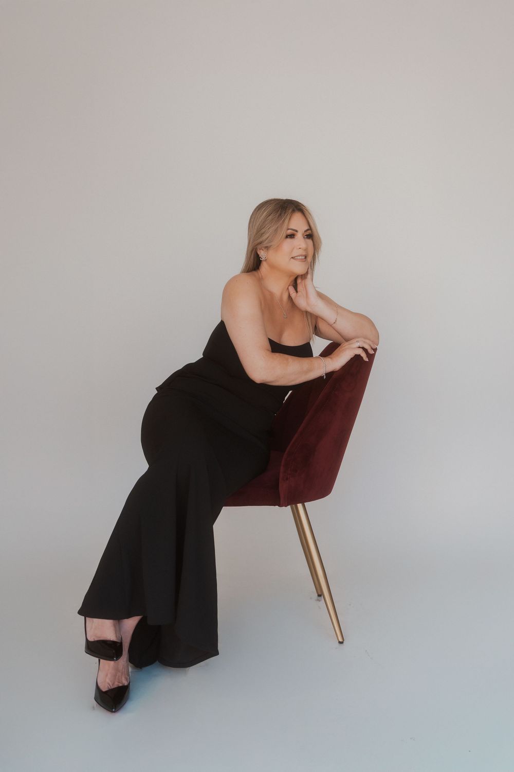 Woman in black evening gown poses elegantly on burgundy chair in studio setting.
