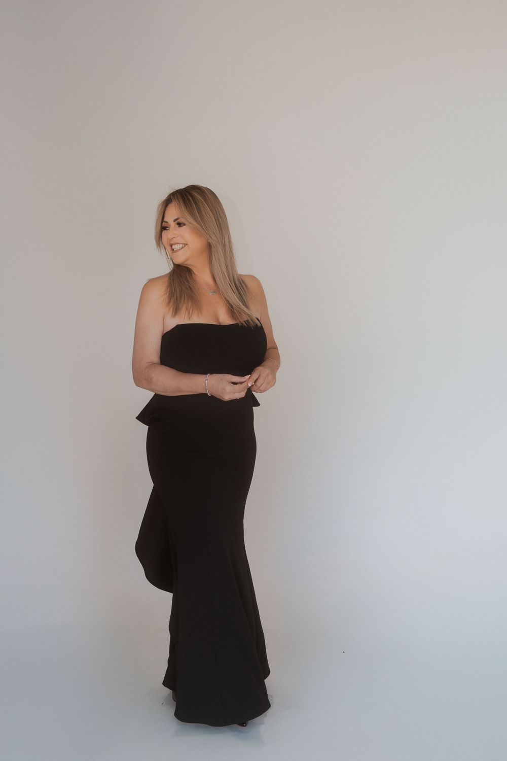 Woman in black formal gown stands confidently against light grey studio background.