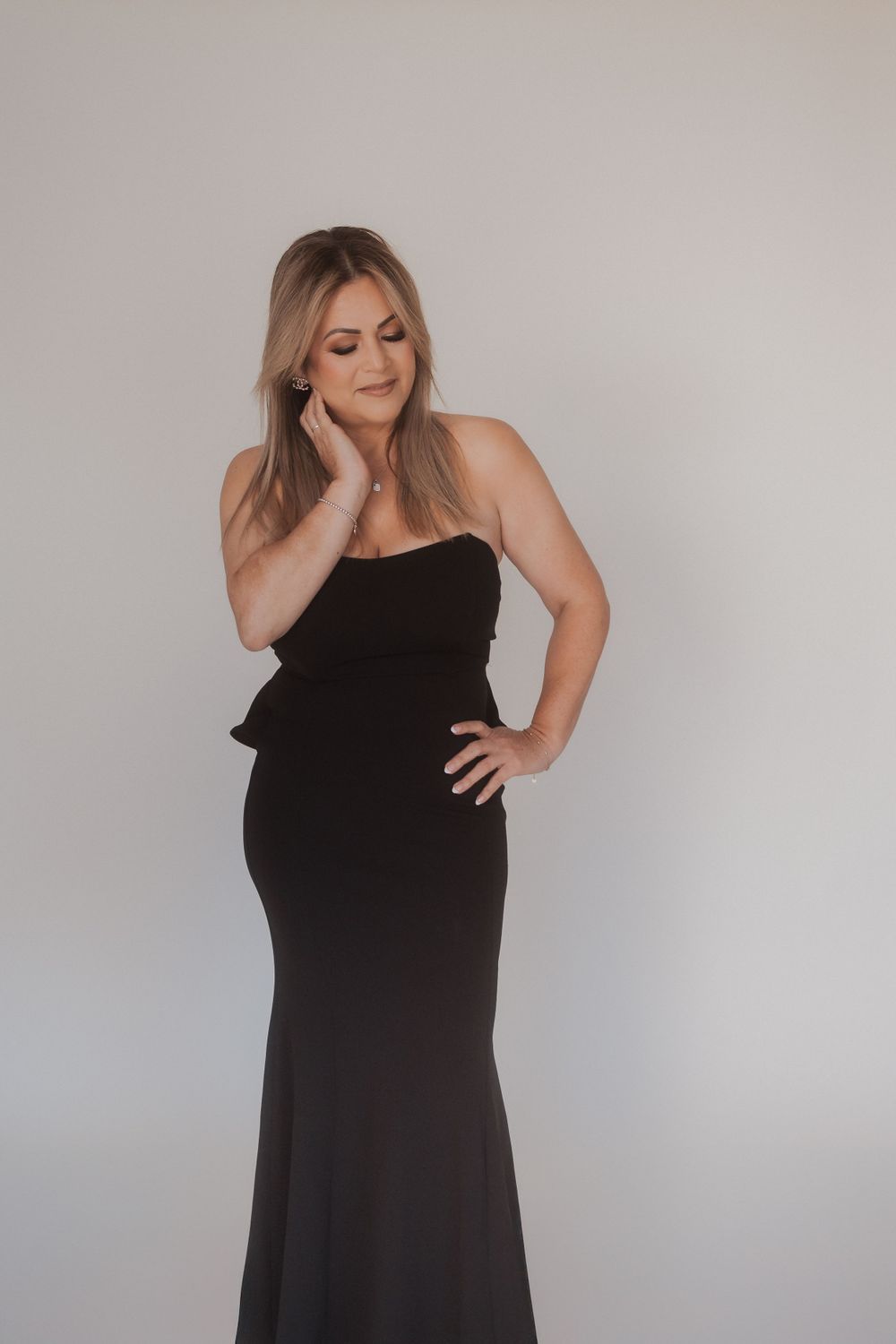 Woman in elegant black strapless evening gown poses against neutral background during professional photoshoot.