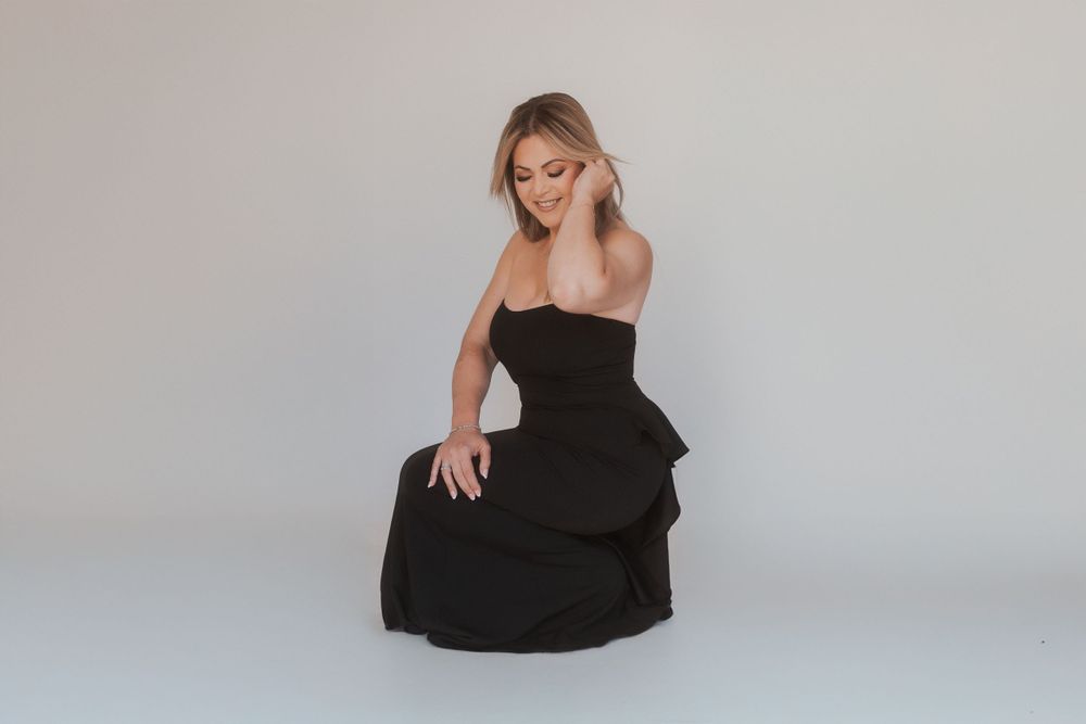 Stylish woman wearing black strapless gown poses during studio photoshoot on light background.