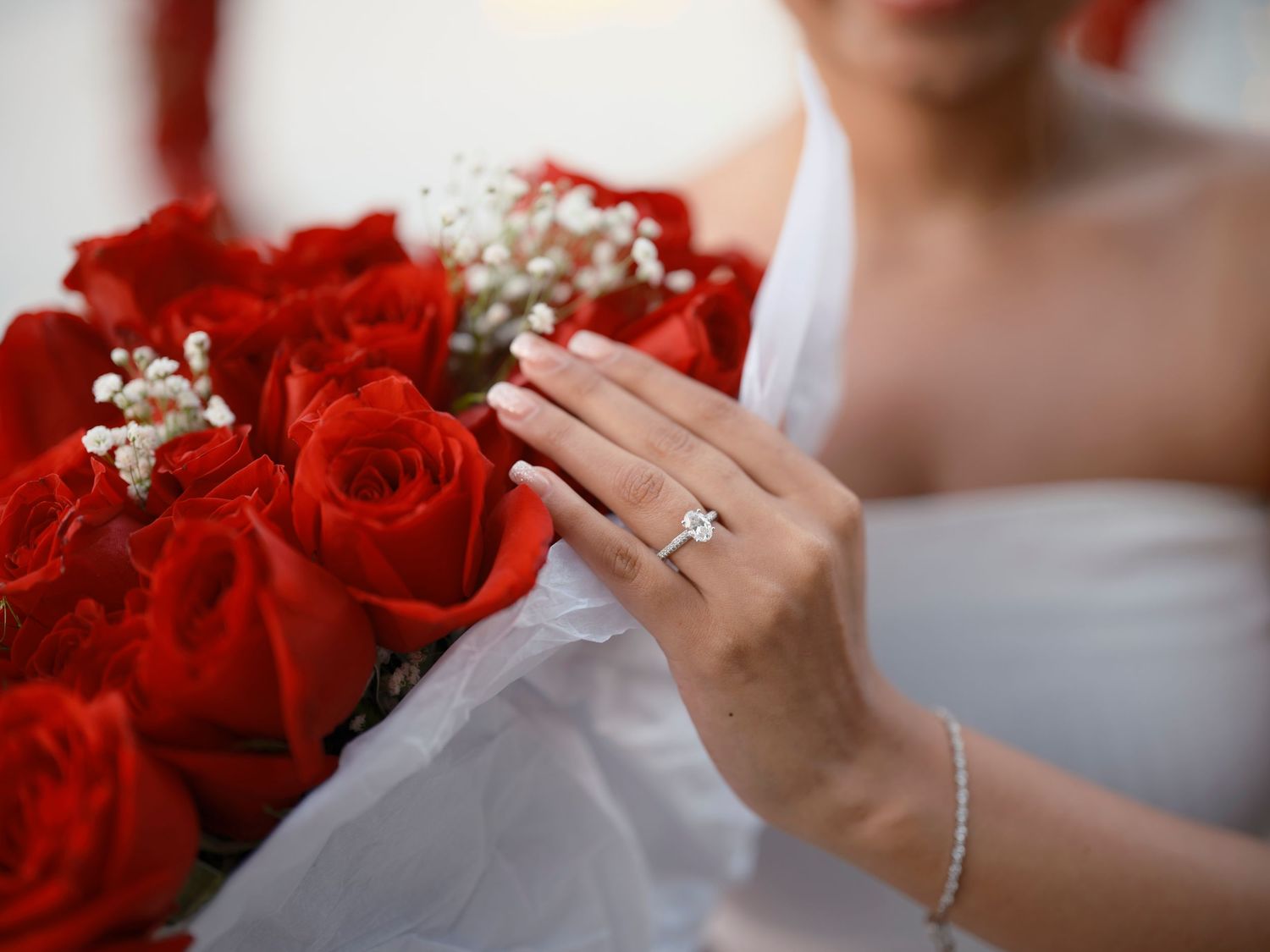 Hands holding red rose bouquet with baby's breath flowers and sparkling engagement ring in romantic wedding setting.