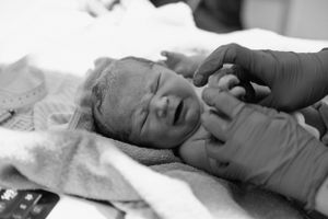 A black and white photograph of a gloved hand gently touching a newborn baby lying on hospital bedding. Birth photography, Karratha, WA