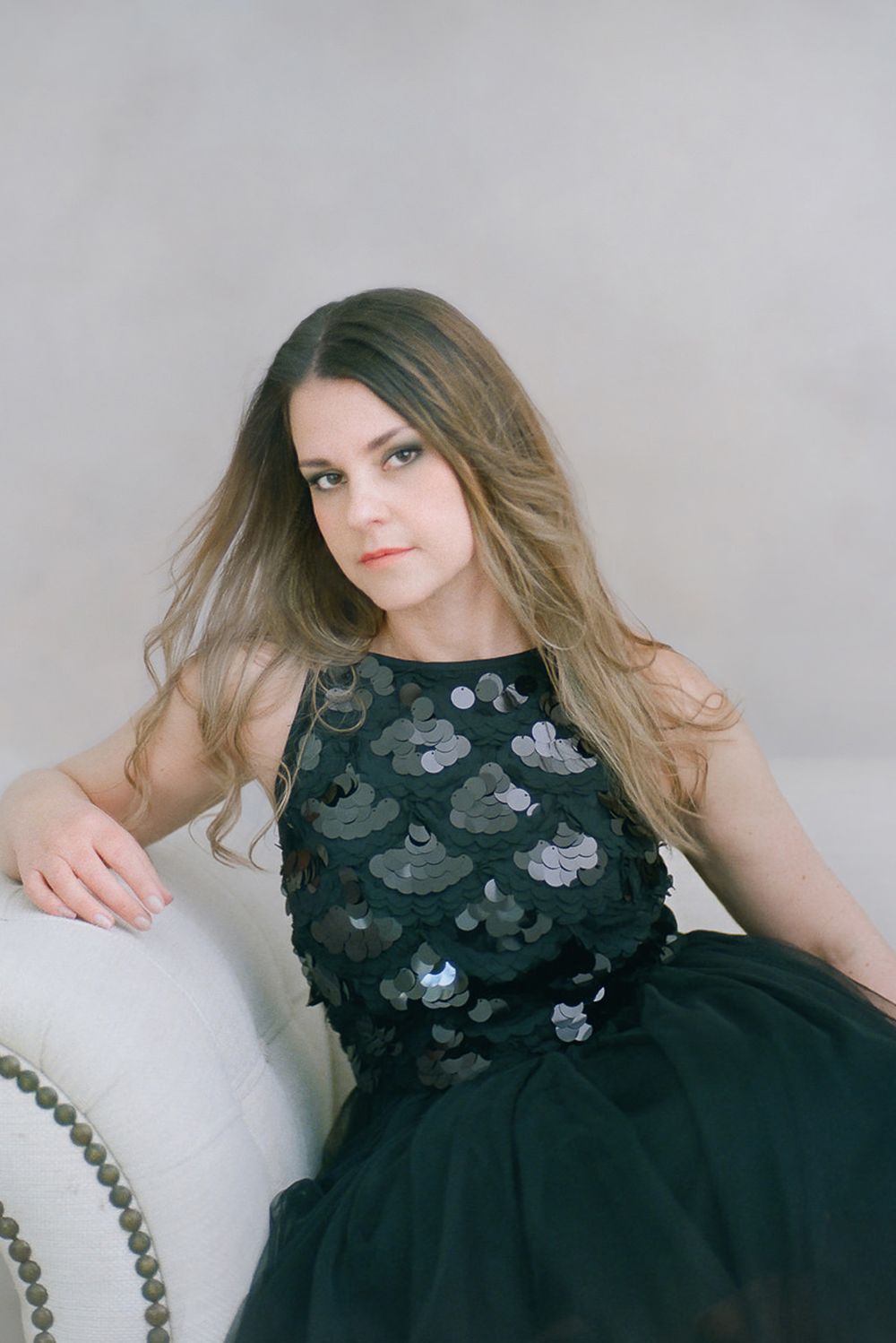 Portrait of person in dark sequined formal dress with floral appliques and tulle skirt reclining against light background.