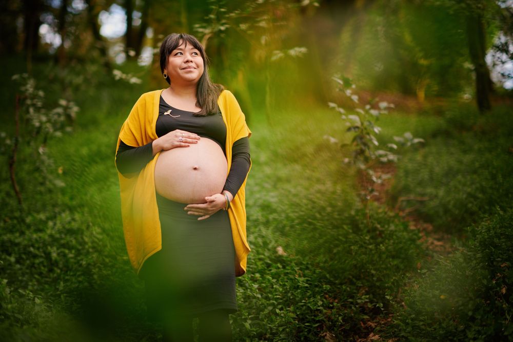 Smiling pregnant woman in yellow cardigan cradling her belly in a sunlit green forest.