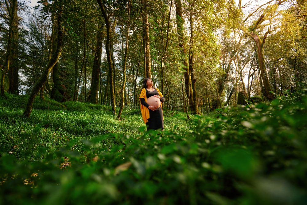 Pregnant woman standing in a lush green forest clearing in a yellow cardigan.