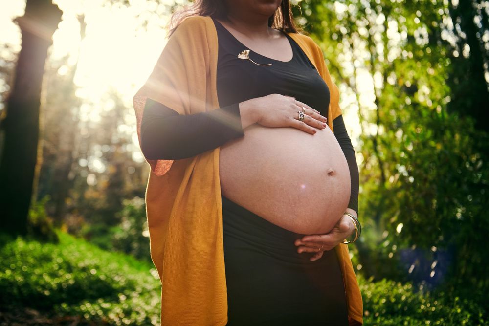 Close-up of a pregnant woman in a yellow cardigan cradling her bare belly outdoors.