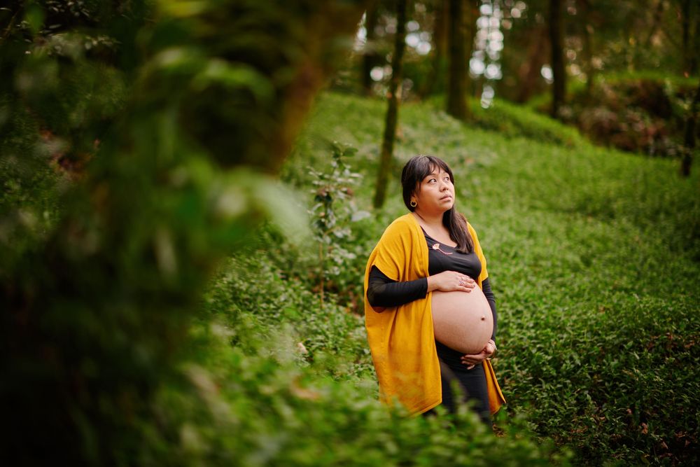 Pregnant woman in yellow cardigan holding her belly while standing in a green forest.
