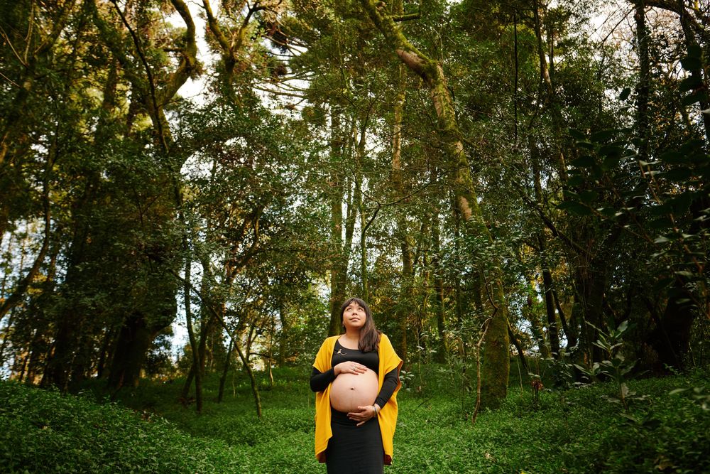 Pregnant woman in yellow cardigan looking up while standing in a lush green forest.