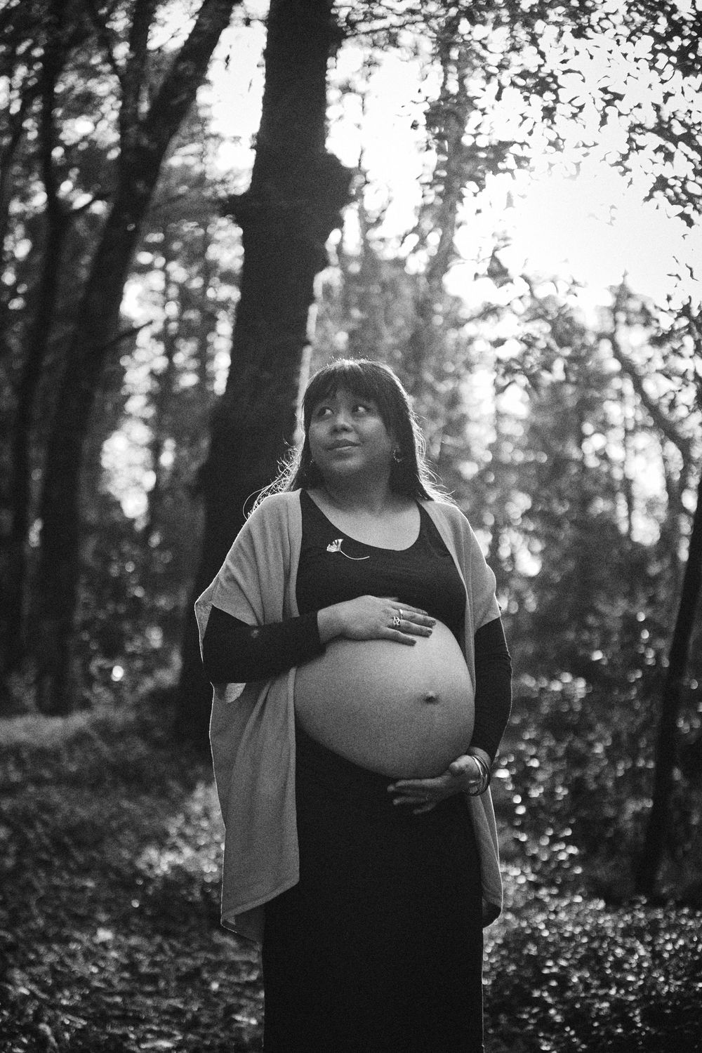 Black and white portrait of a pregnant woman holding her bare belly in a forest.