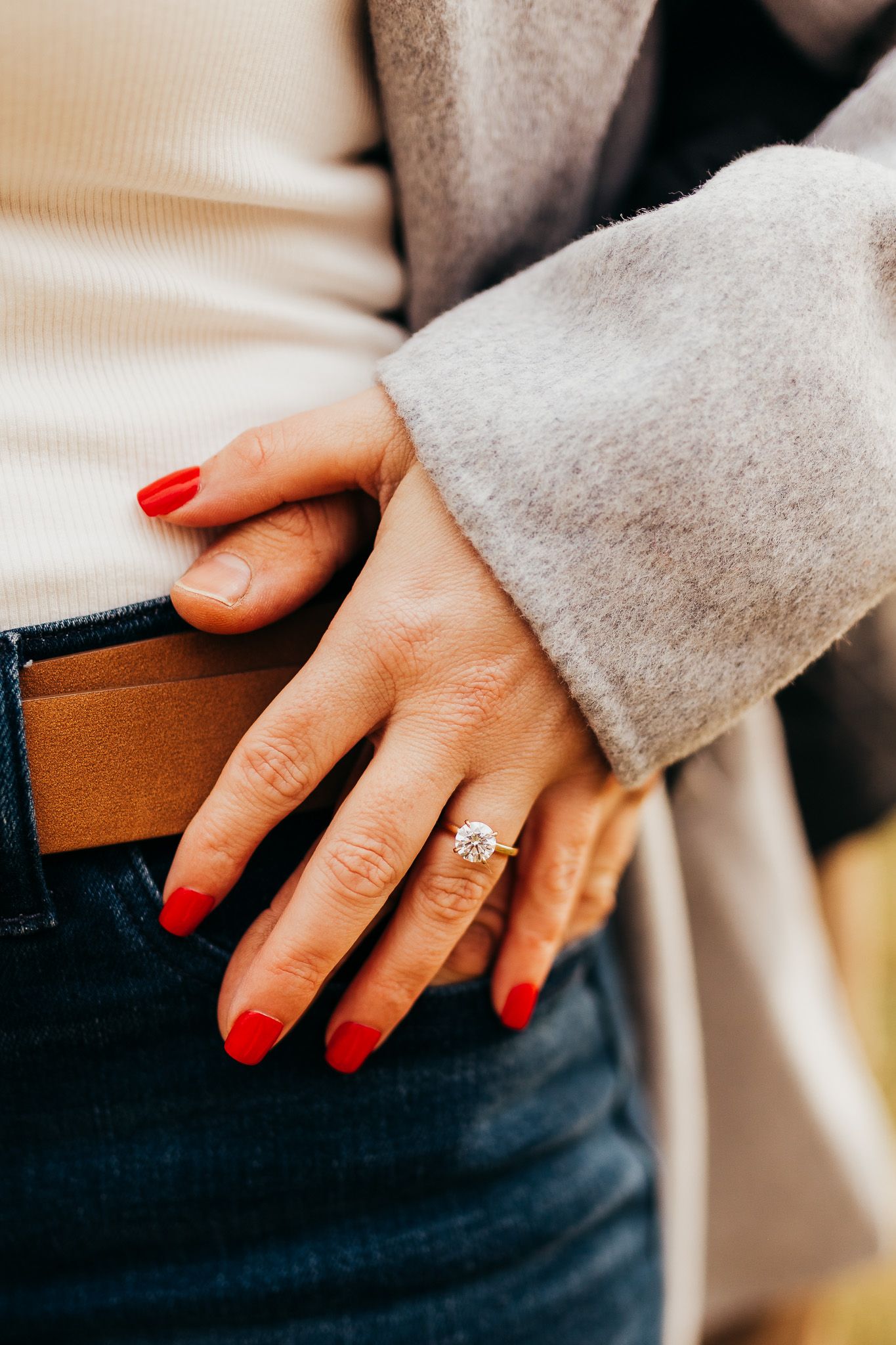 Close-up of hand wearing engagement ring with red nail polish against grey sweater.