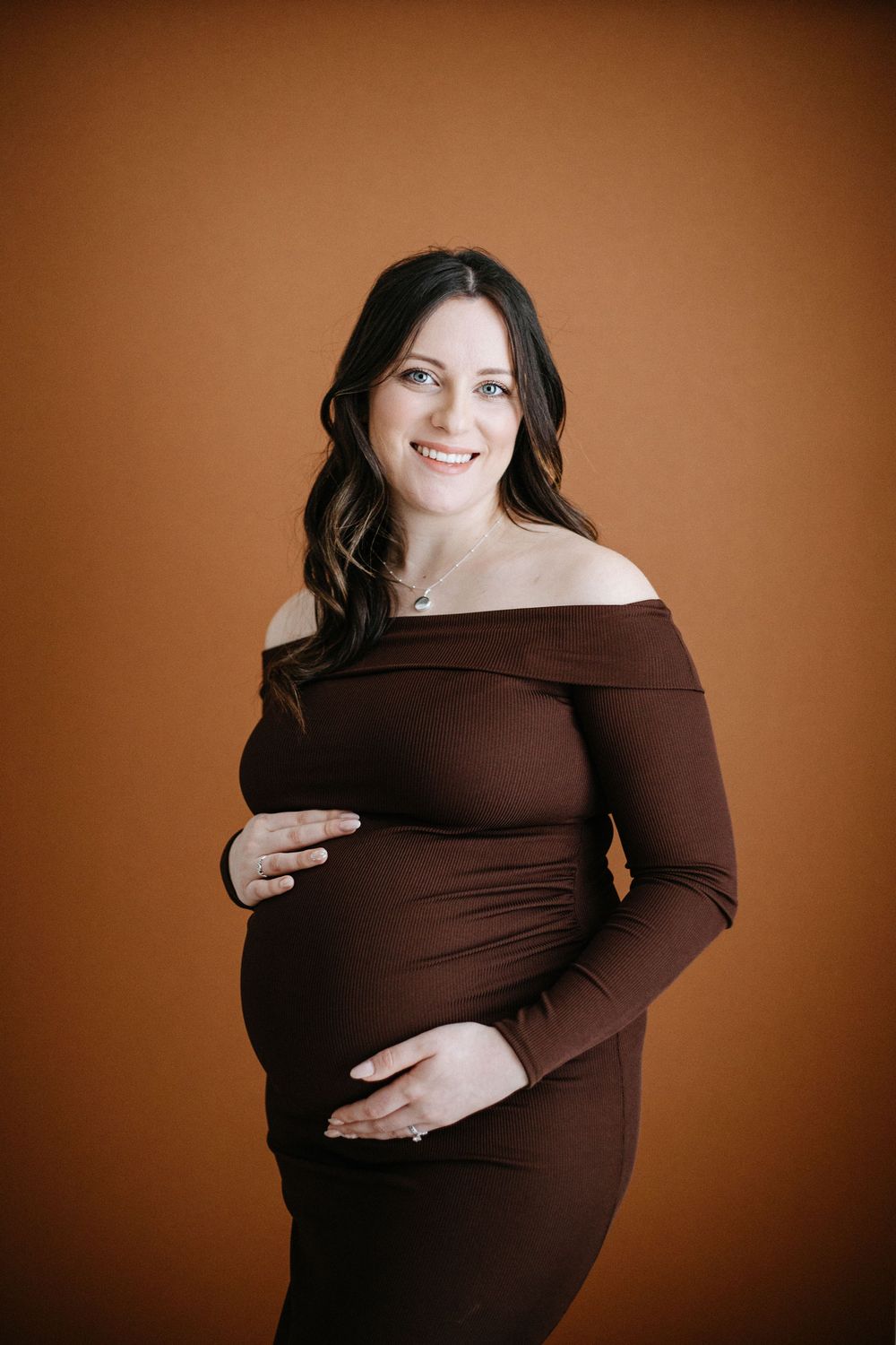 A pregnant woman in a brown off-shoulder dress poses against a warm brown background during a maternity photoshoot.