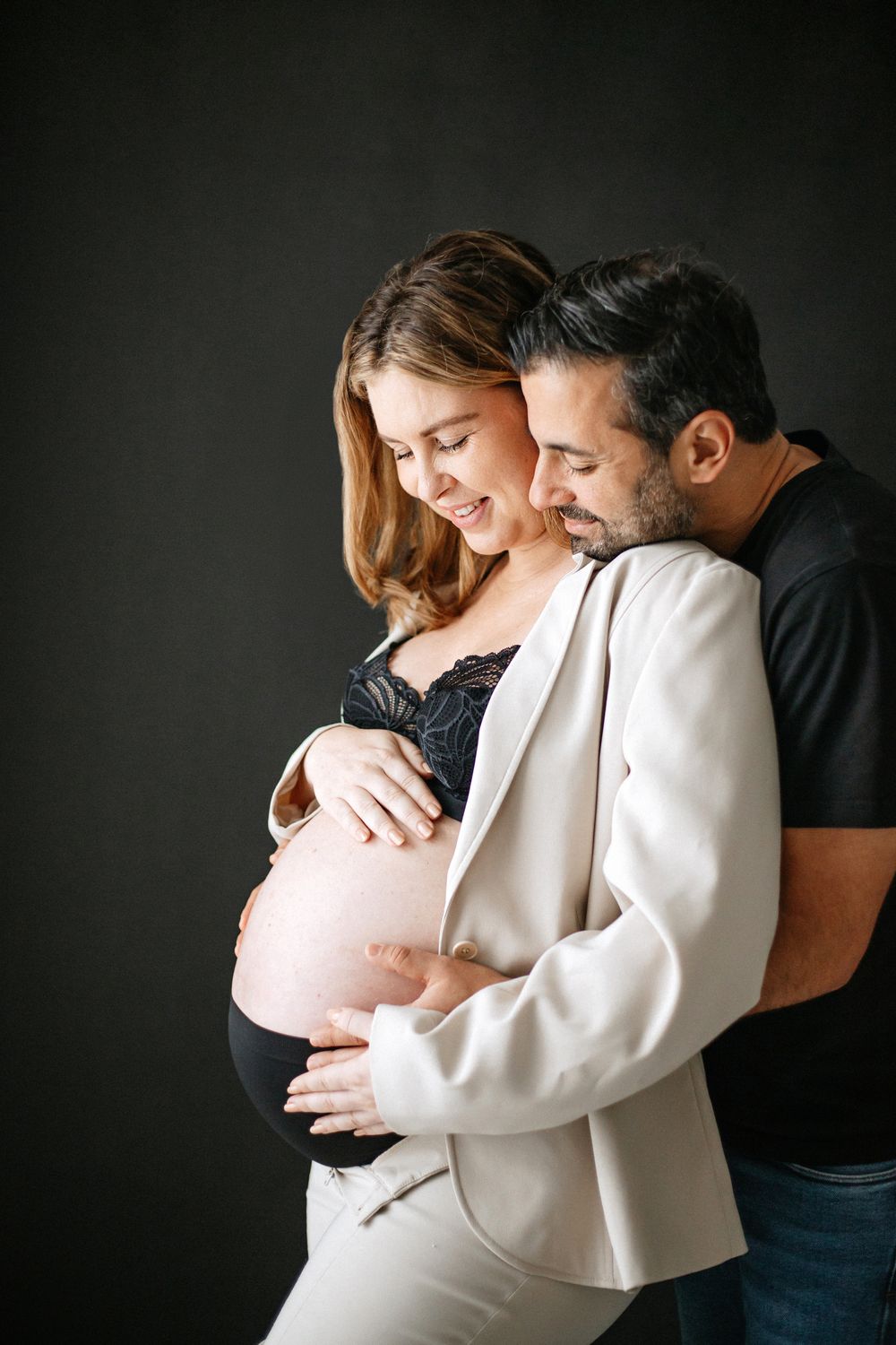 Intimate maternity photo shows expectant couple embracing against dark background in modern studio setting.