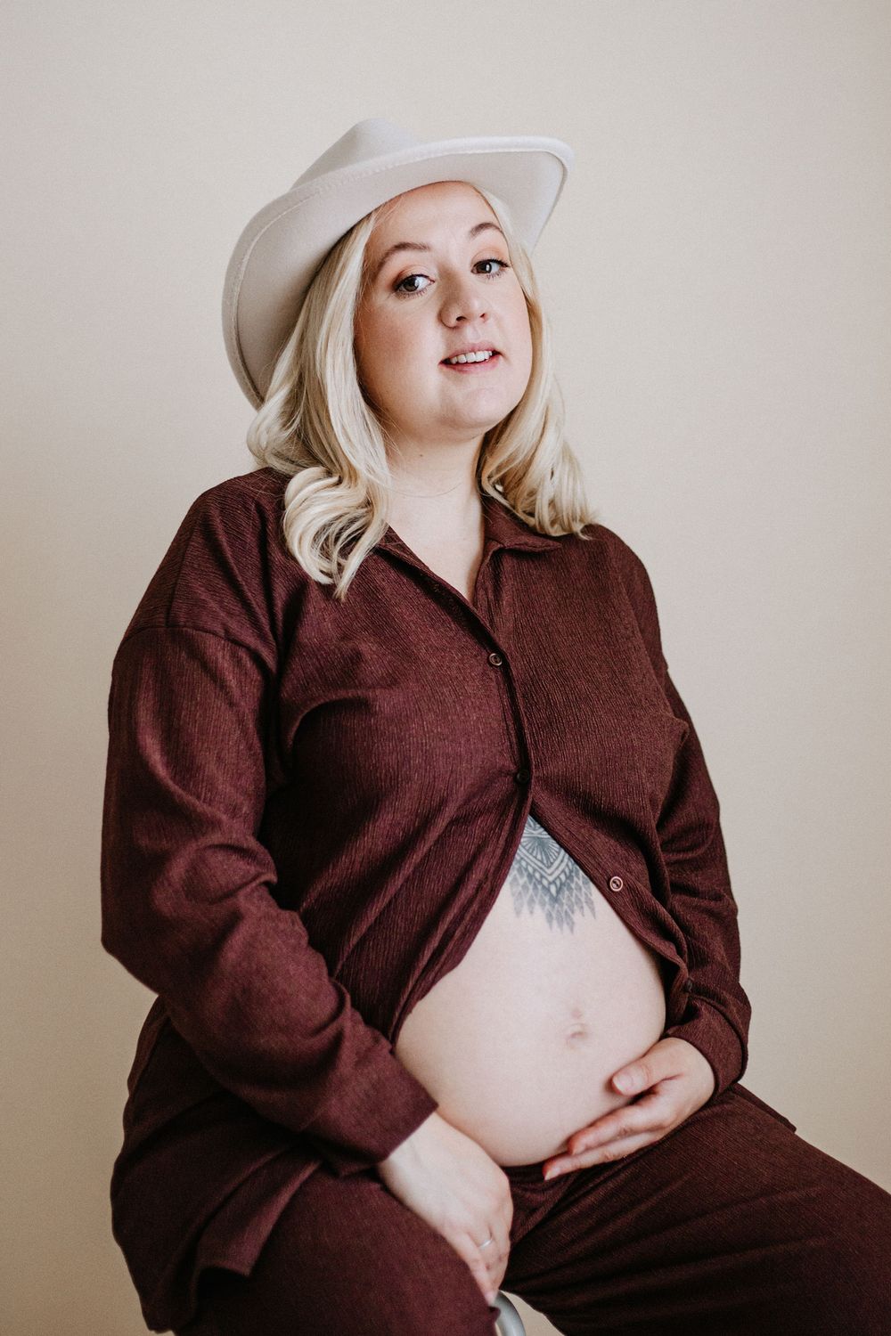 A pregnant person wearing a brown outfit and white hat poses against a neutral background.