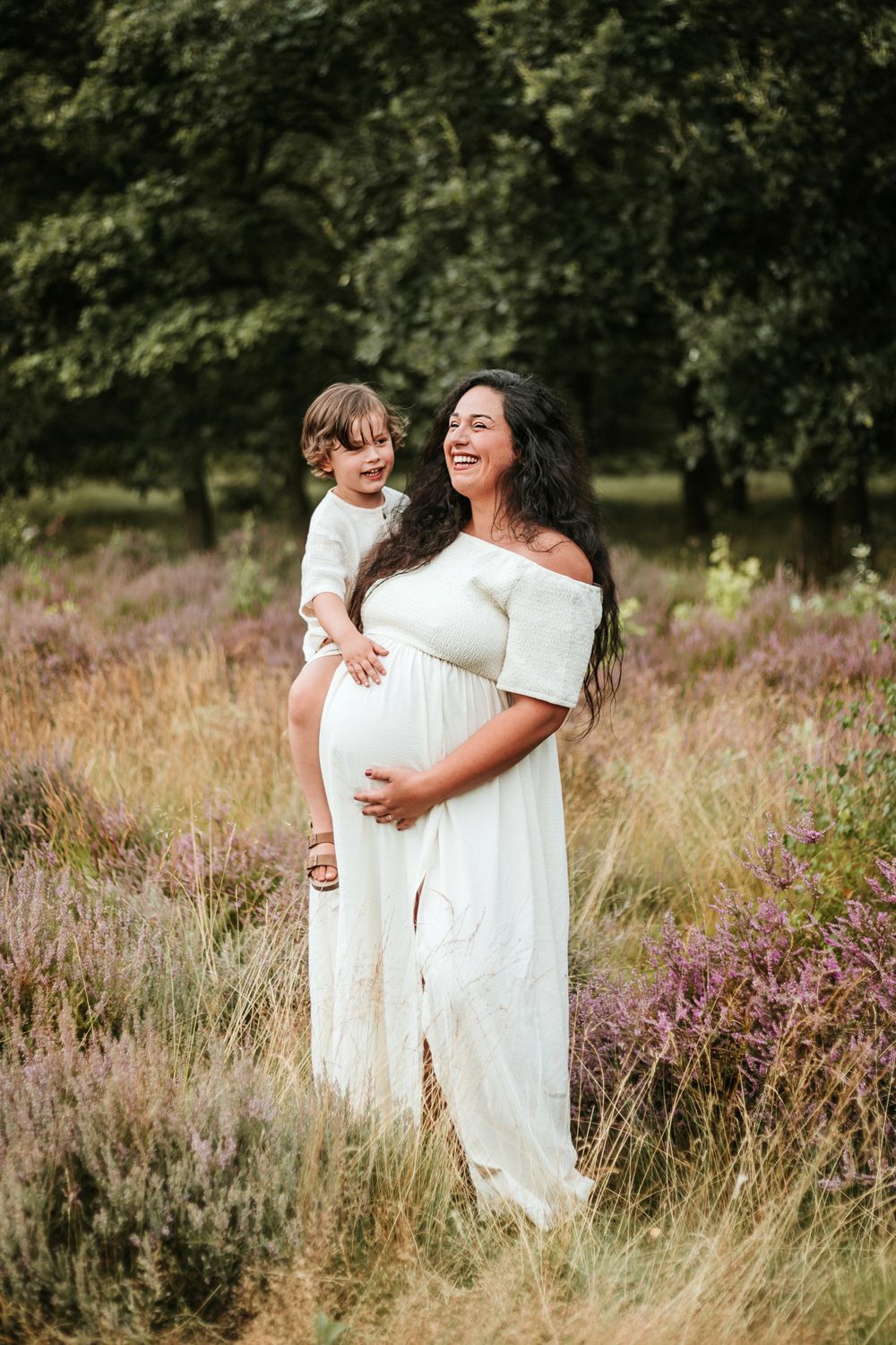 A pregnant woman in a white dress stands in a meadow of wildflowers and tall grass during golden hour.