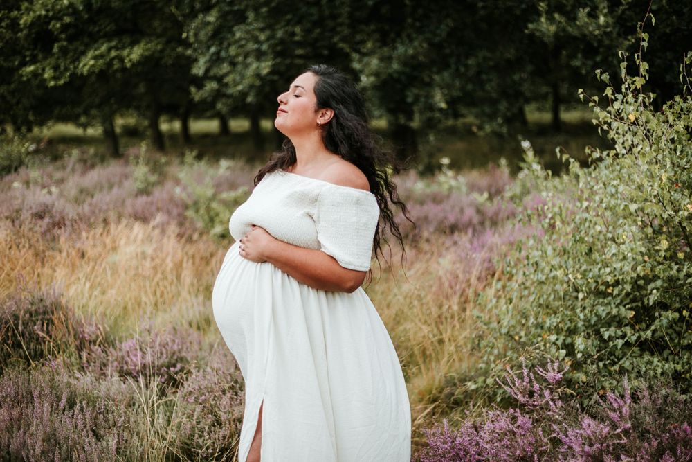 A person in a white maternity dress stands in a field of heather and wildflowers during sunset.