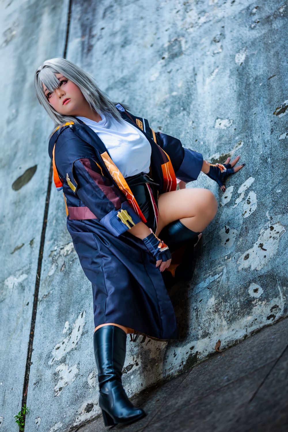 A cosplayer in a navy uniform and silver wig poses against a concrete wall.