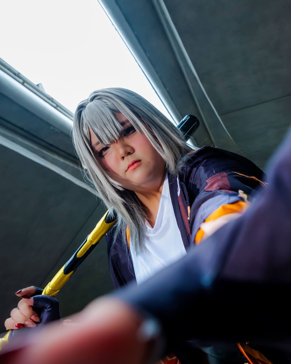 A cosplayer wearing a white and navy outfit poses with dramatic lighting against a dark background.
