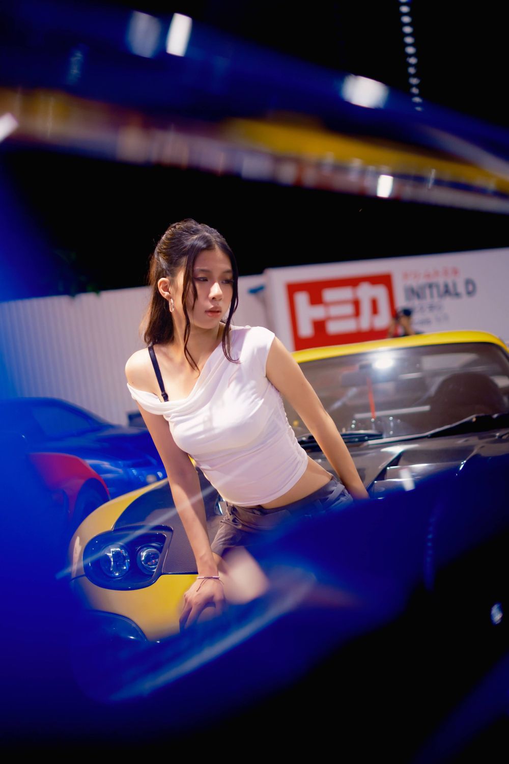 Model in white crop top poses with toy car at automotive event display.