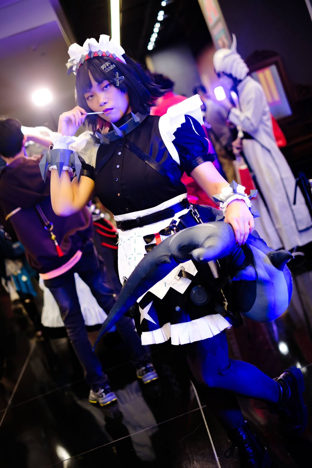 A cosplayer in a navy and white maid uniform performs an energetic dance pose on stage.