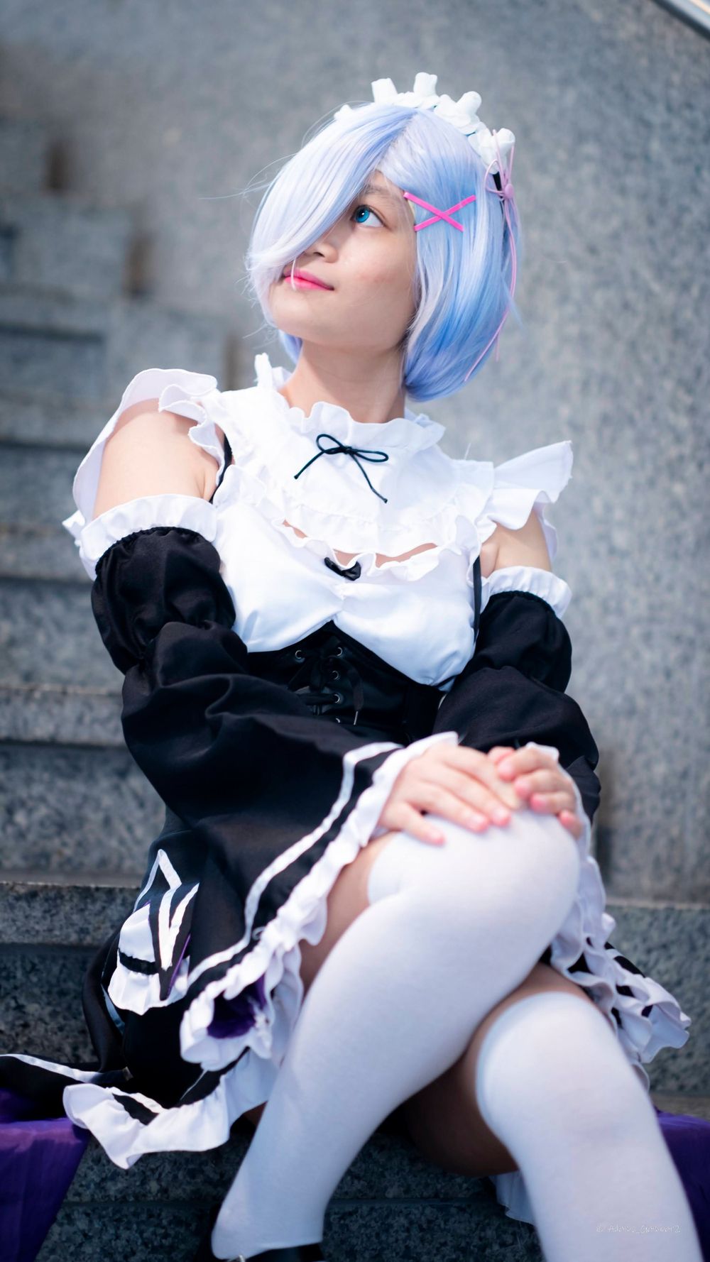 A cosplayer in a maid outfit with blue and white hair poses on concrete steps.