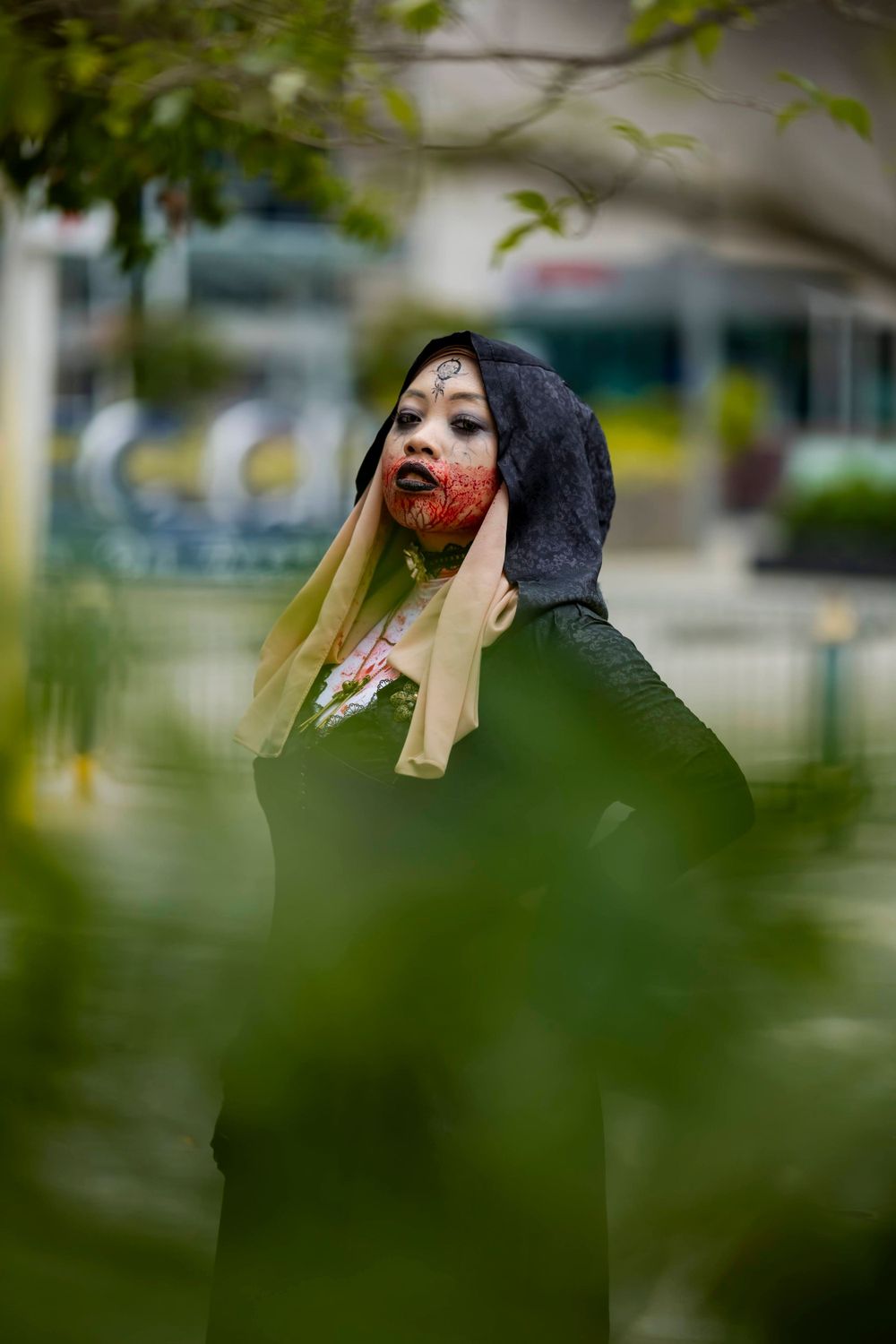 A figure in a black hooded cloak with dramatic makeup poses among green foliage.