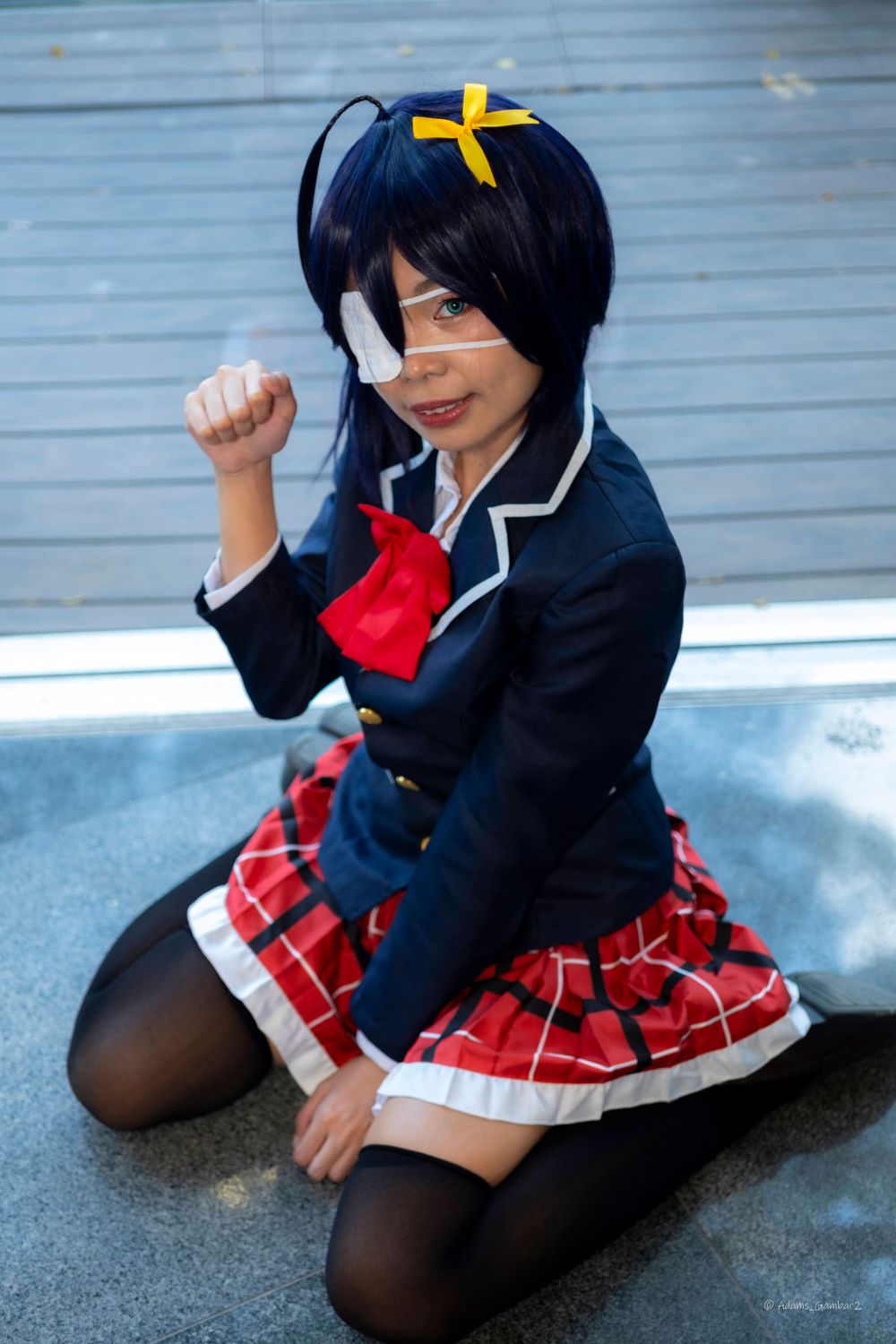 A cosplayer wearing a navy uniform and plaid skirt poses on the ground.