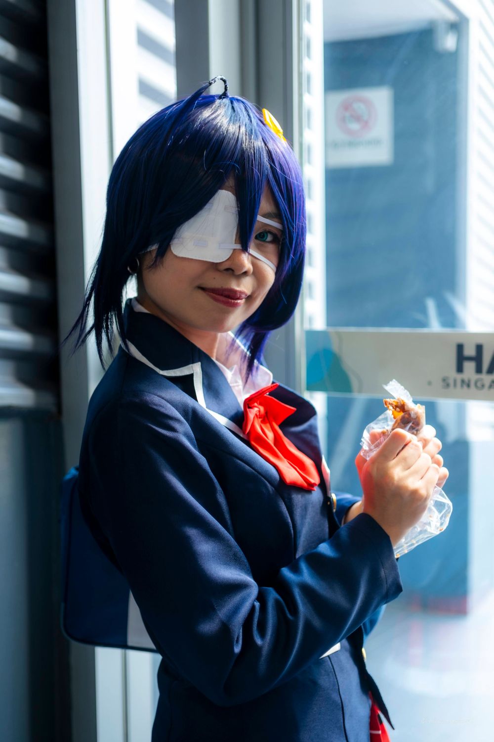 A cosplayer in a navy school uniform with blue hair poses against shutters.