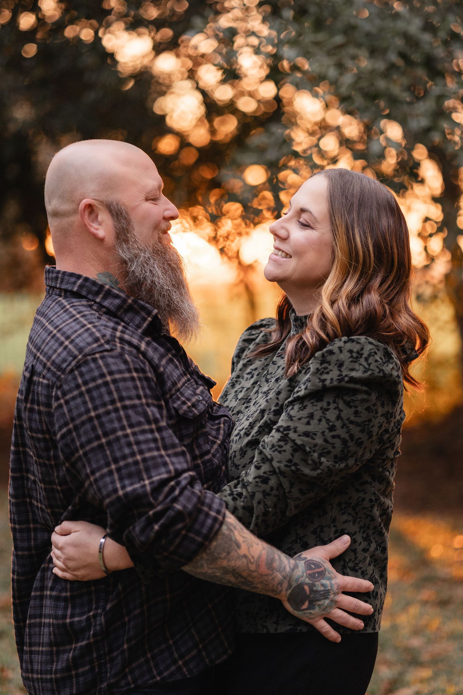 Couple embracing outdoors at golden hour, bald bearded tattooed man and smiling brunette woman in autumn park setting.