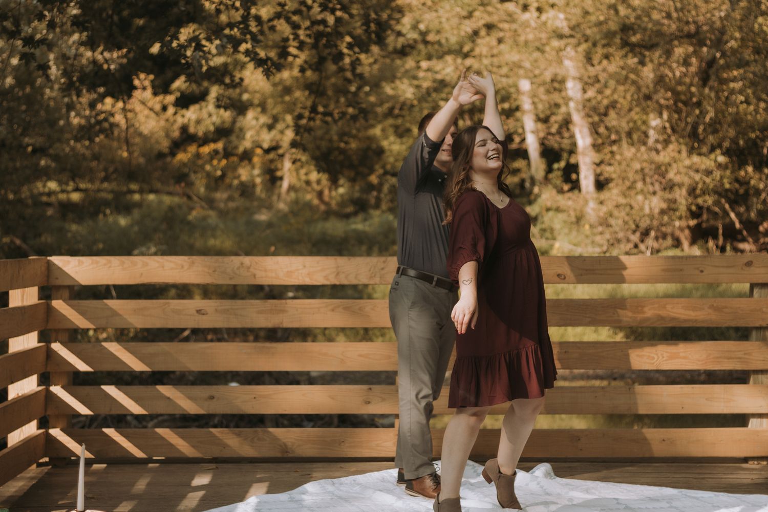 A couple in autumn outfits stand at a wooden railing during a fall engagement photoshoot with golden leaves in background.