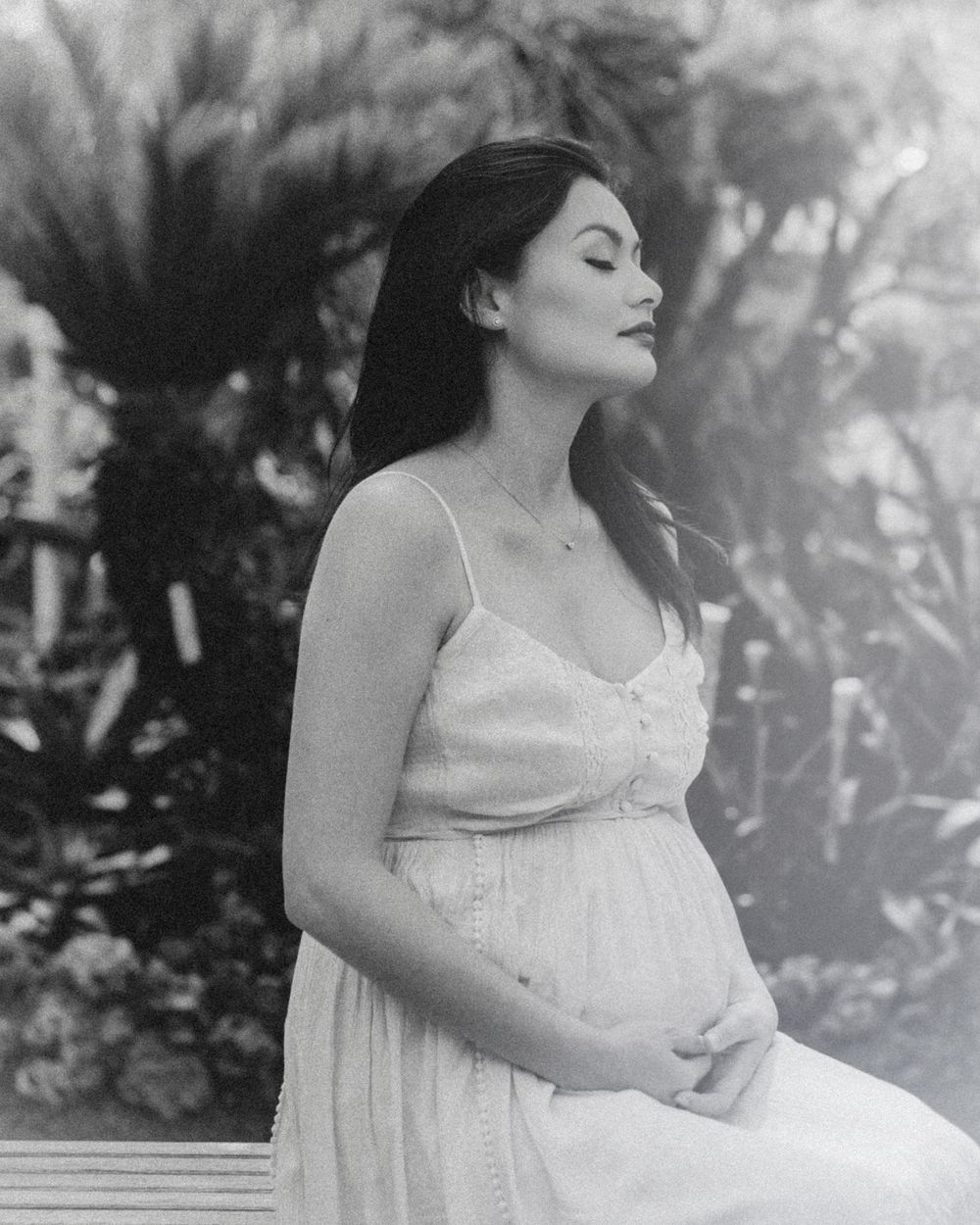 Serene black and white maternity portrait in a dress against lush garden backdrop.
