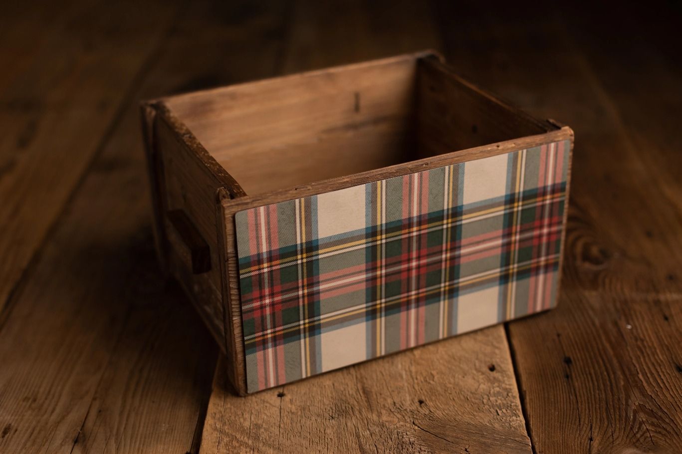 A rustic wooden crate decorated with tartan plaid fabric trim in red, green, and cream on a dark wood floor.