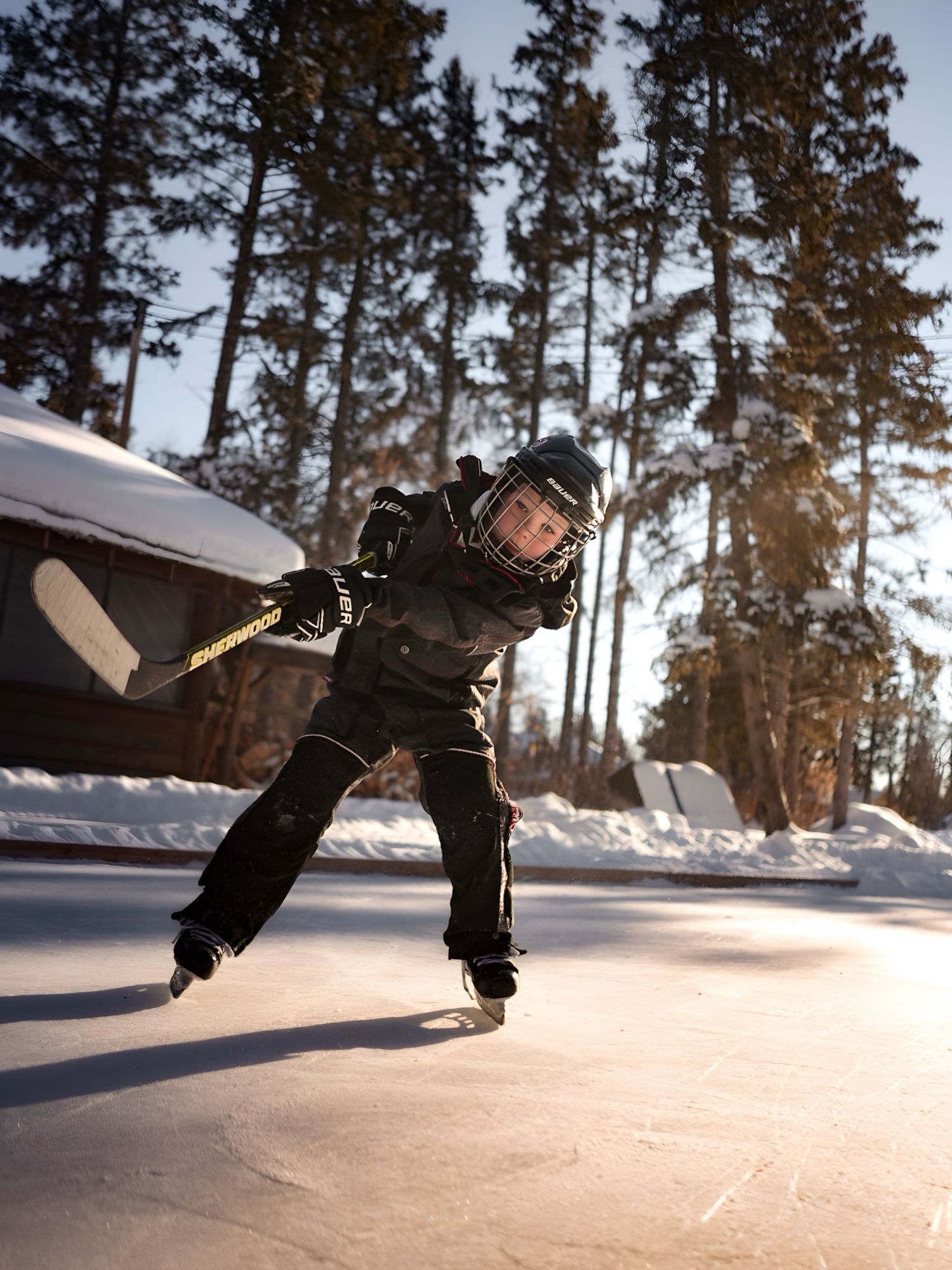 Ice skater performing jumps on a frozen outdoor rink surrounded by snow and pine trees.