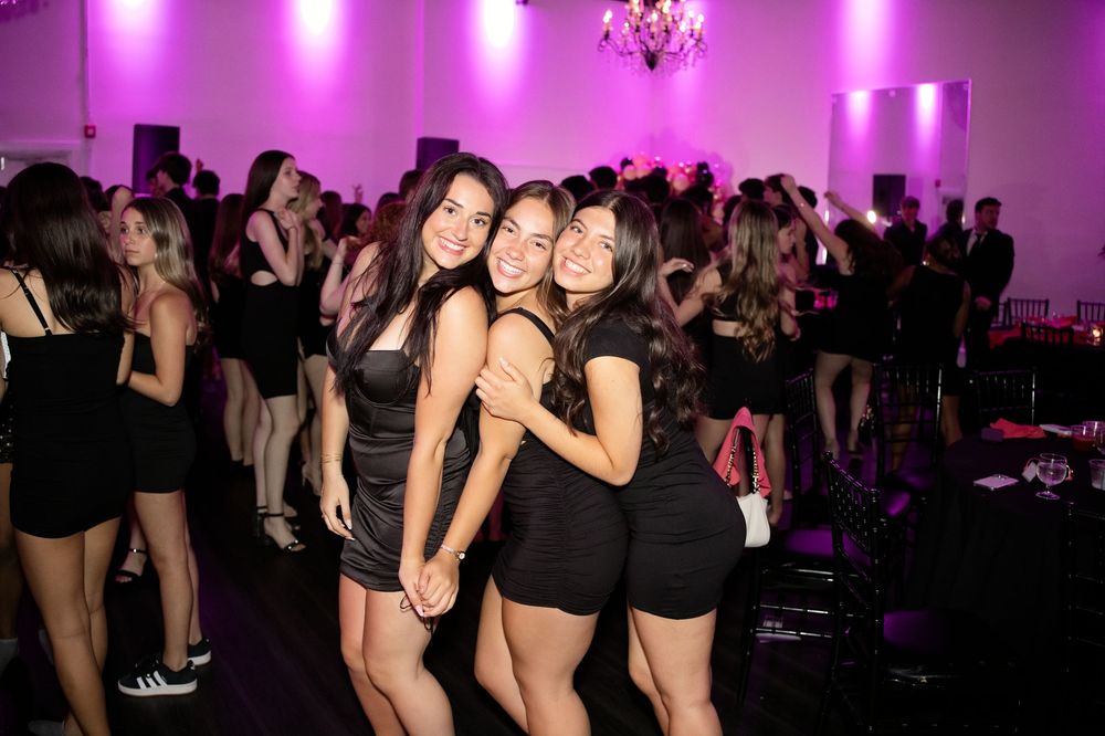 Three young women in black outfits smile and pose together at a lively evening party with pink lighting.