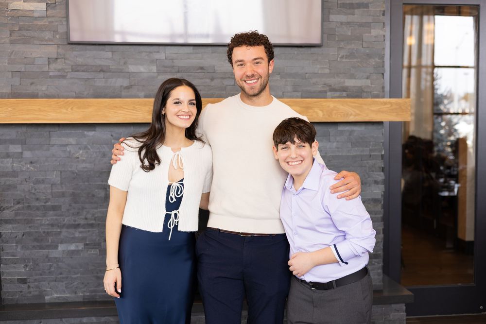 Three people smiling and posing together in front of a stone wall backdrop at a celebration event.