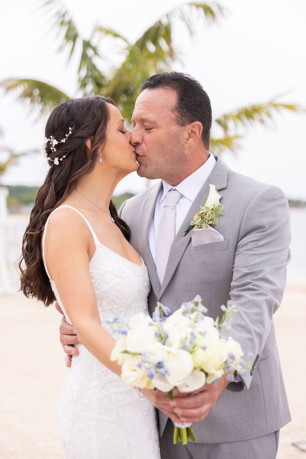 Newlyweds share a romantic kiss on the beach, bride holding a white and blue floral bouquet.
