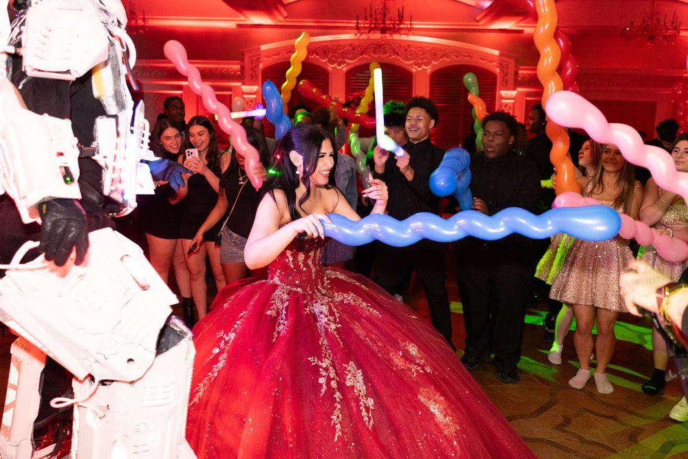 Young woman in red ball gown celebrates quinceañera surrounded by guests holding colorful balloon sculptures.