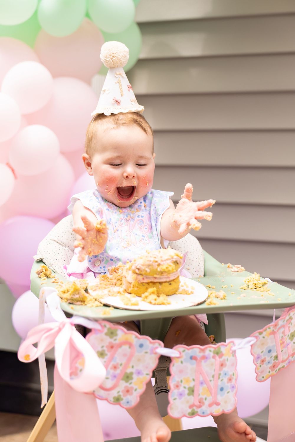 Baby in party hat joyfully smashing first birthday cake in high chair, surrounded by pink and green balloons.