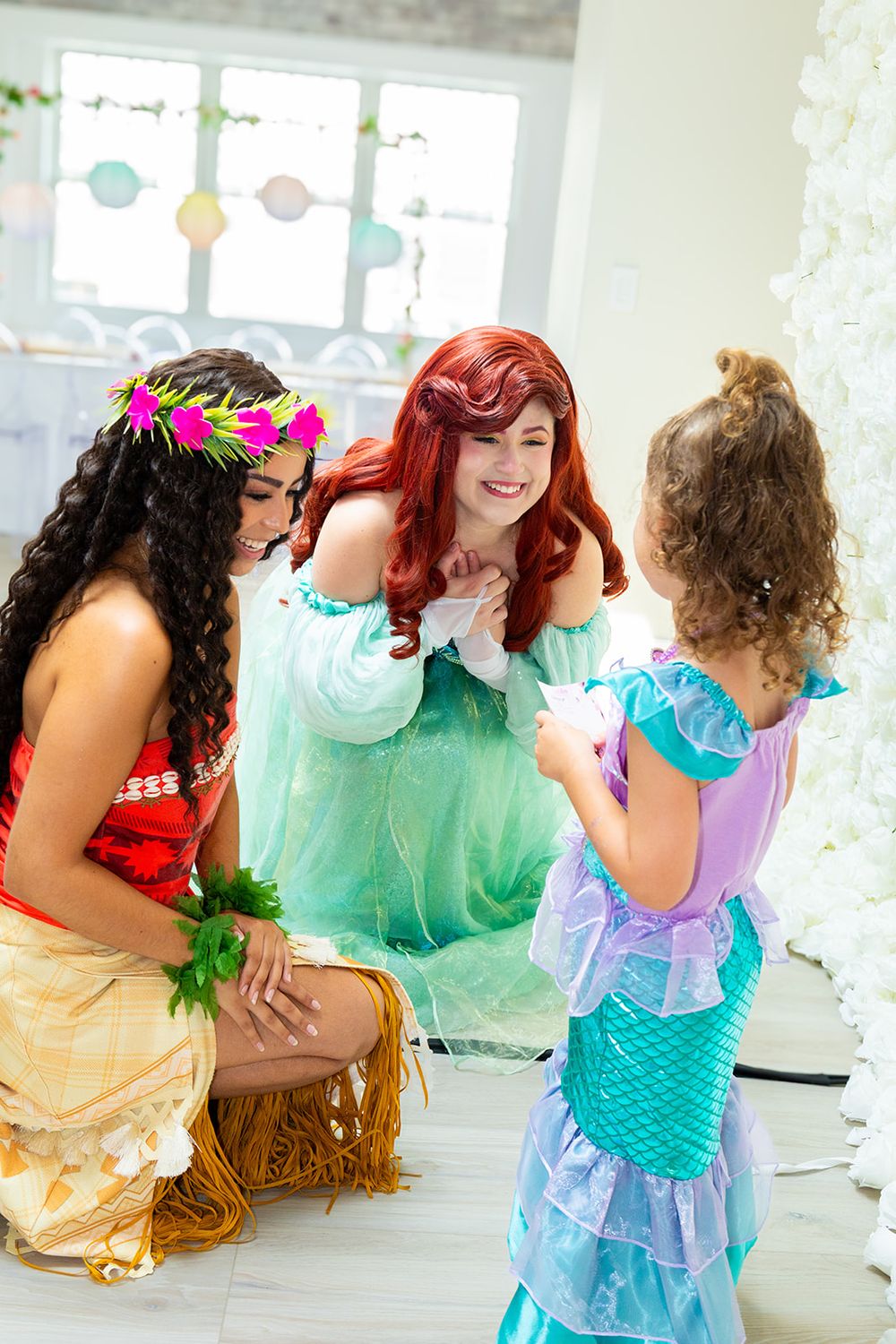 Two women dressed as Disney princesses Moana and Ariel interacting with a young girl.