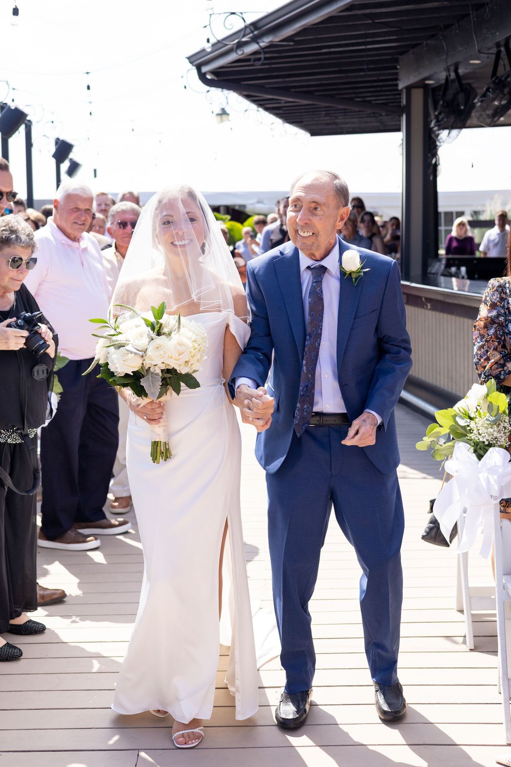 Bride in white gown and veil walks down aisle escorted by older man in navy suit at outdoor wedding.