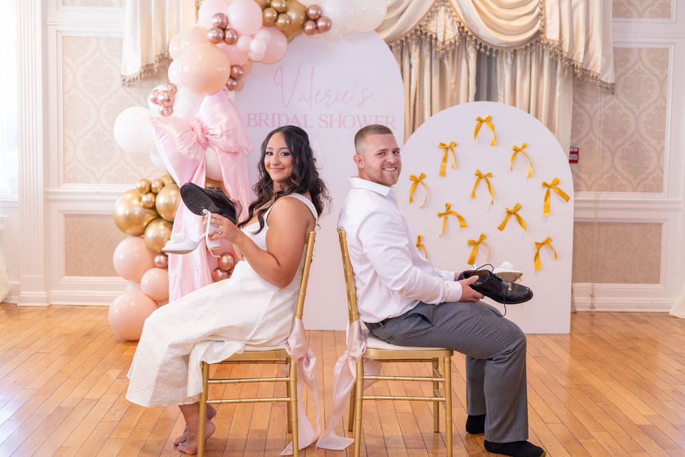 A couple sits back-to-back at a baby shower gender reveal, holding pink and black baby shoes near balloons.