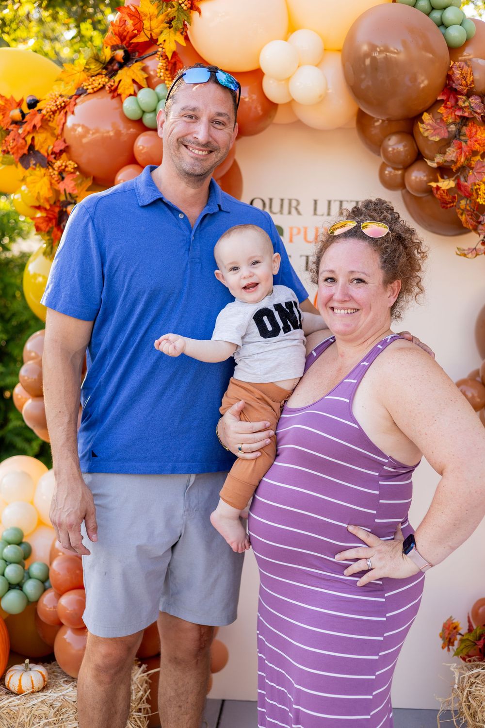 Smiling family with baby in 'ONE' shirt at fall-themed first birthday party with balloon arch.
