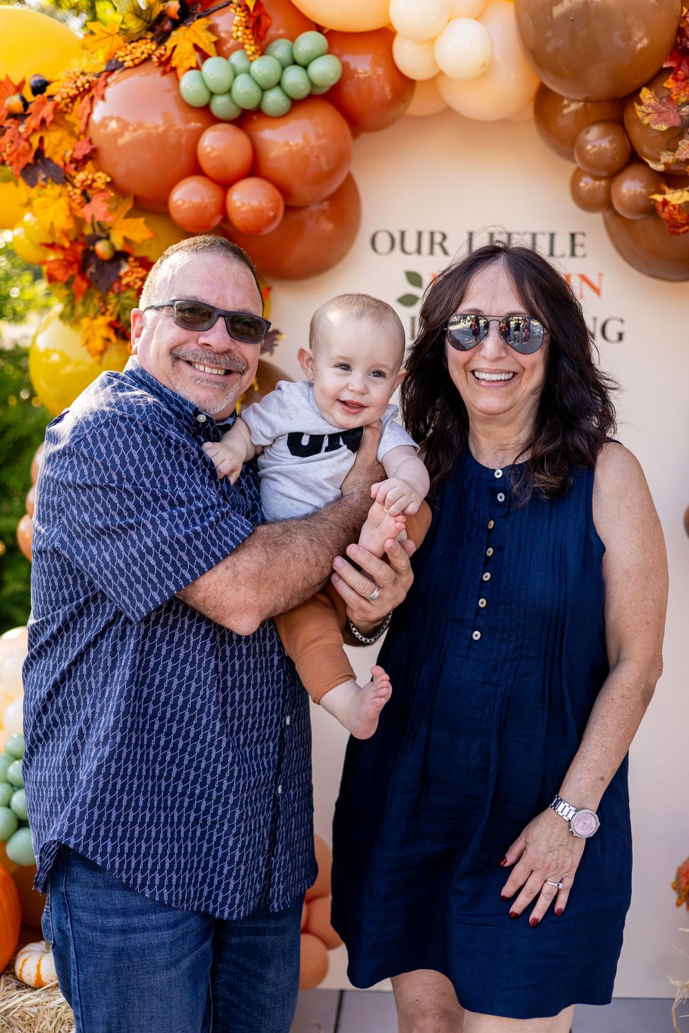 Grandparents smiling and holding a happy baby at a fall-themed first birthday party with balloon arch.