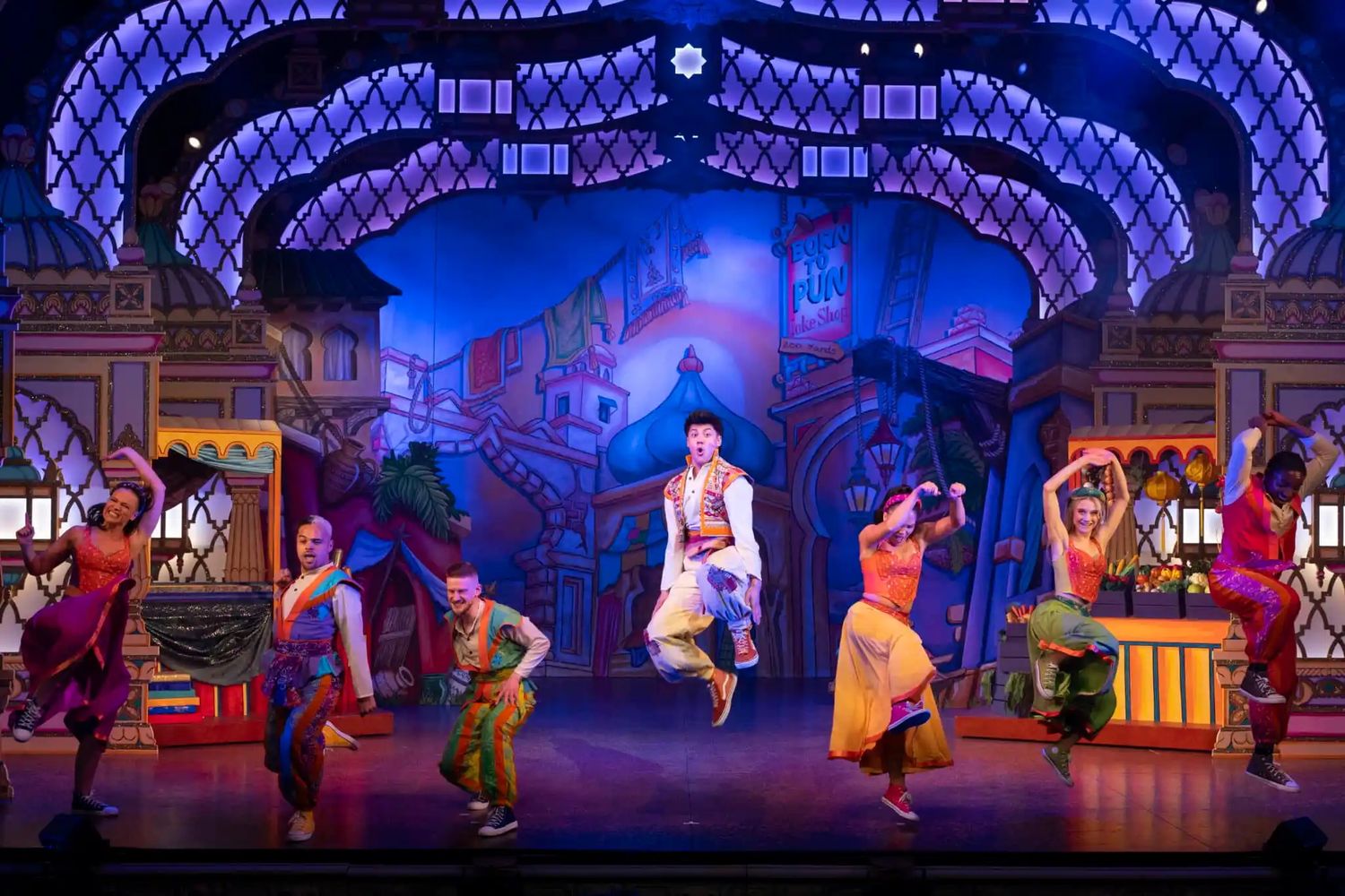 Dynamic stage performance with dancers in period costumes beneath blue-lit latticed ceiling decorations.